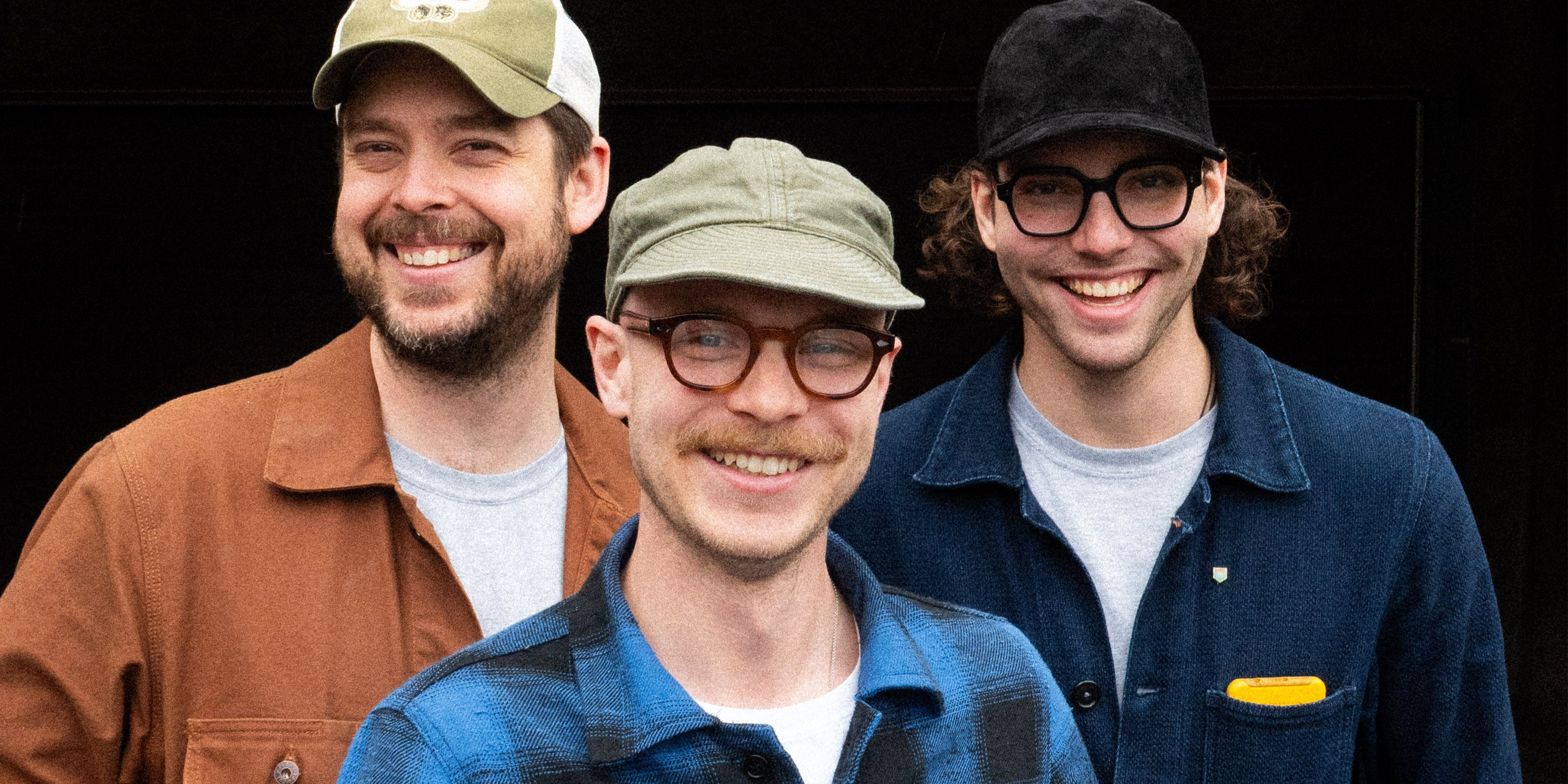 Three men wearing hats and smiling against a black background