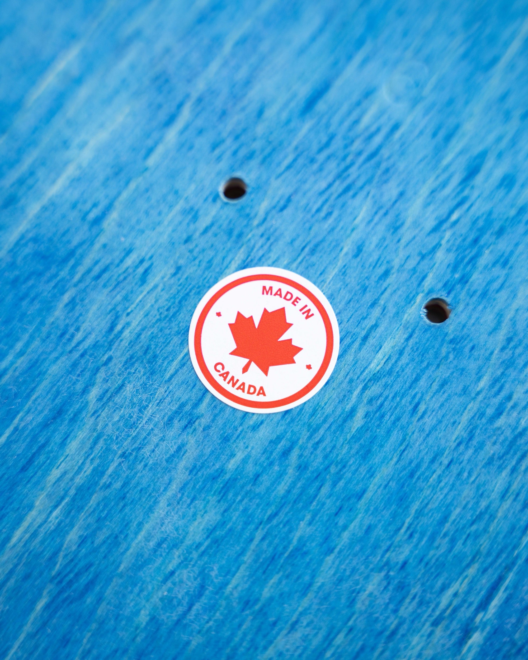 A circular red and white 'MADE IN CANADA' sticker with a maple leaf design, shown on a blue wood-grain surface.