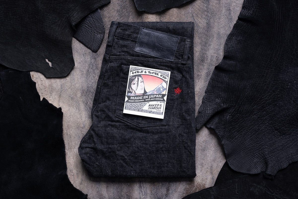 A pair of folded black denim jeans with a paper brand label on the back pocket, arranged on a textured gray surface surrounded by dark fabric pieces. The label reads 'Mats & More Co' and 'Made in Japan'.